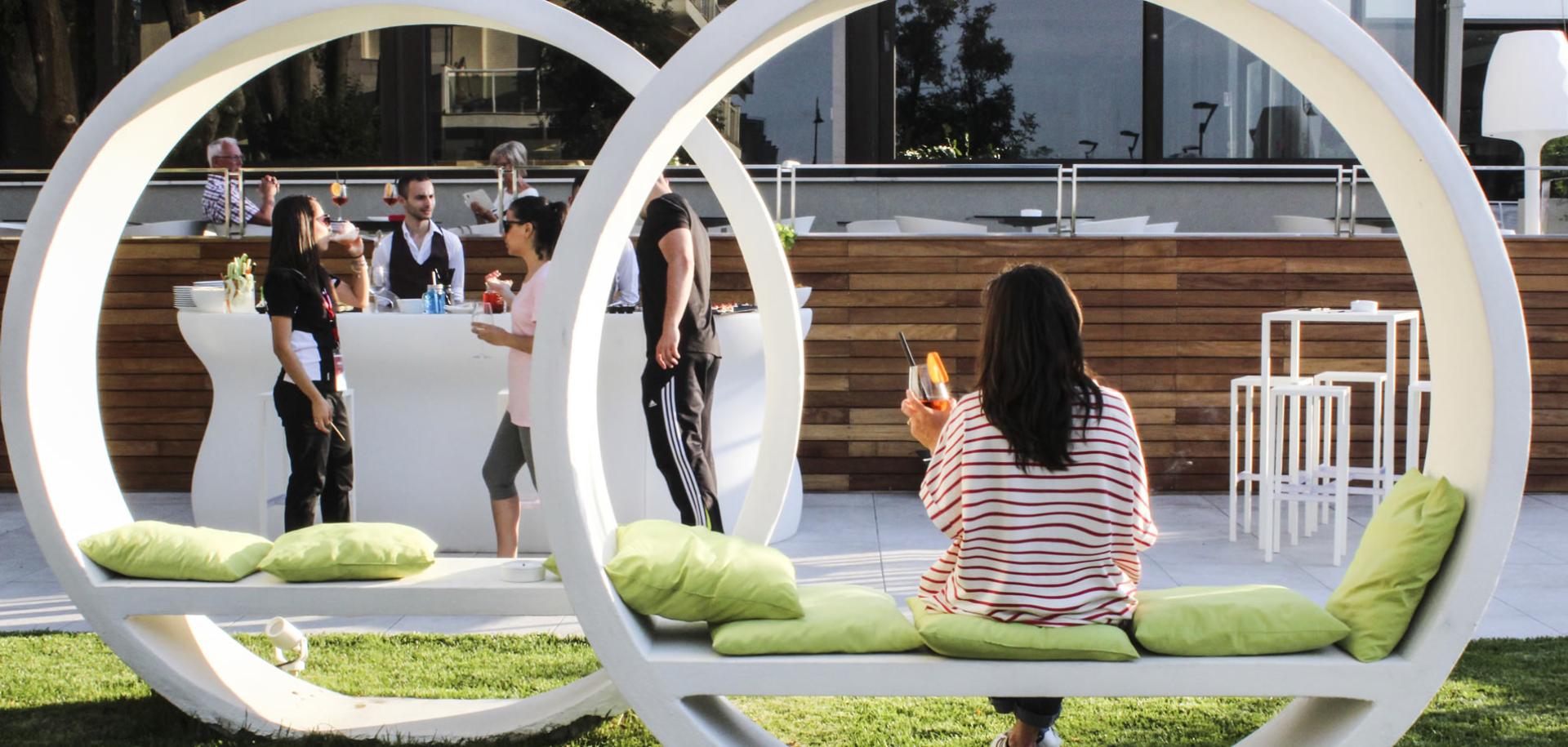People socializing outdoors with drinks, sitting on modern circular chairs.